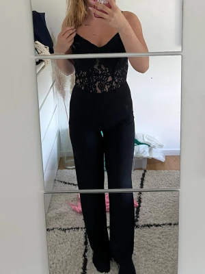 Svart spetsjumpsuit med smala axelband - Säljer en svart jumpsuit med smala axelband och v-ringning. Överdelen är i genombruten spets med snygg detalj på ryggen och dragkedja. Byxdelen är lång och rak. Perfekt för dig som vill ha en stilren och trendig look.