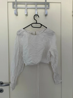 Vit croppad blus med knyt - Trendig vit croppad blus med långa puffärmar och smock vid ärmsluten. Blusen har rund halsringning och öppen rygg med knytband i midjan. Perfekt för en stilren och modern look.