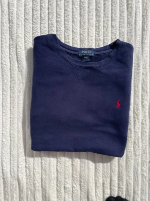 Mörkblå sweatshirt från Polo Ralph Lauren - Klassisk mörkblå sweatshirt från Polo Ralph Lauren med rund halsringning och den ikoniska röda logotypen broderad på bröstet. Tröjan har långa ärmar och ribbade muddar vid ärmslut och nederkant. Perfekt för en avslappnad och stilren look.
