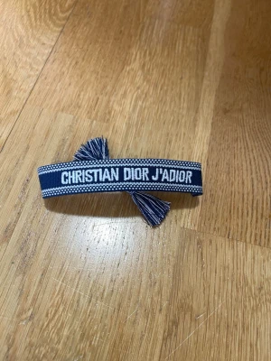 Christian Dior J'Adior blått armband - Snyggt blått tygarmband från Christian Dior med texten 'J'Adior' broderad i vitt. Armbandet har justerbar knytning med tofsar i ändarna och en cool, casual vibe. Perfekt accessoar för att lyfta din outfit med en touch av lyx.