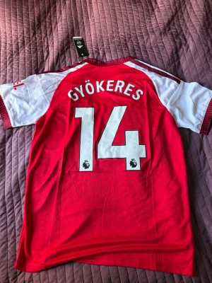Arsenal matchtröja Gyökeres 14 - Röd och vit Arsenal matchtröja från Adidas med Gyökeres och nummer 14 på ryggen. Tröjan har klubbmärke och Adidas-logga på bröstet samt sponsortryck. Tillverkad i lätt och ventilerande material, perfekt för fotboll eller träning.