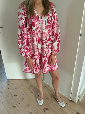 Vit och rosa blommig kortklänning xs - Supersöt kortklänning med volangkant och v-ringning i vitt och rosa med blommigt mönster. Klänningen har långa, puffiga ärmar och en loose passform som ger en avslappnad och trendig look. Perfekt för sommaren!