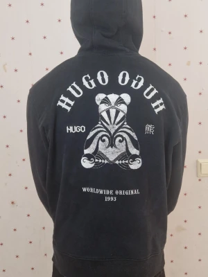 Svart hoodie från Hugo med björntryck - Svart hoodie från Hugo med stor vit grafisk björn och text på ryggen. Framtill finns en liten björnlogga på bröstet och en klassisk magficka. Huva med dragsko och ribbade muddar. Perfekt för dig som gillar streetwear och unika prints.