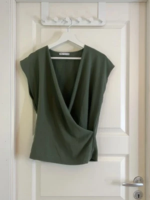 Olivgrön omlottopp från Zara - Snygg olivgrön omlottopp från Zara med djup v-ringning och ärmlös design. Toppen har en avslappnad passform och är tillverkad i mjukt bomullsmaterial. Perfekt att styla med jeans eller kjol för en trendig look.