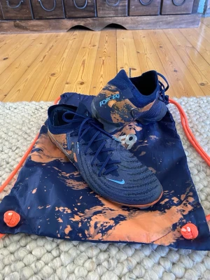 Nike phantom gx2 Haaaland edition  - Säljer ett par Nike fotbollsskor i blått med orange detaljer och mönster.  Änvönda i en säsong, ogroligt sköna och passa främst för medel/ lite bredare fötter. Medföljer matchande blå och orange gympåse och kartong.