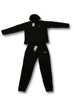 Nike Miller  - Nike Träningsset / Tracksuit – Hoodjacka + Byxor – Helt Ny med Taggar! Snyggt helsvart Nike träningsset bestående av en lätt hooded zip-up jacka och matchande byxor. Officiellt Nike-set i perfekt nyskick, aldrig använt och med alla originaltaggar kvar. Detaljer: •  Märke: Nike •  Storlek: M (Medium) •  Färg: Svart •  Setet innehåller: Hoodjacka + byxor •  Egenskaper: Lätt material, elastisk midja och muddar, Nike Swoosh-logga •  Skick: Ny med taggar (deadstock) •  Stil: Sportig / casual tränings