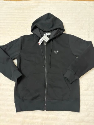 Svart hoodie från Comme des Garçons Play - Svart zip-hoodie från Comme des Garçons Play med huva och dragsko. Fram finns ett litet hjärta med ögon på bröstet och på ryggen ett större hjärta med ögon. Mjuk bomullskvalitet och två fickor framtill. Perfekt för en stilren och cool look.