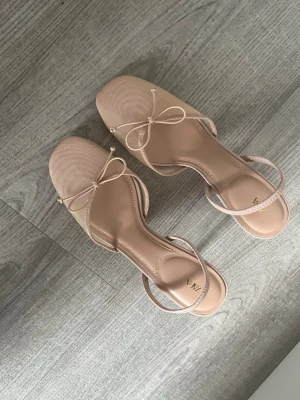 Beige ballerinaskor från Zara - Snygga ballerinaskor från Zara i beige med tunn rem runt hälen och dekorativ rosett framtill. Skorna har en mjuk, rundad tå och är tillverkade i ett tygmaterial som ger en stilren look. Perfekta för dig som gillar minimalistisk och trendig stil. Dem är i storlek 39, men känns som 38! 