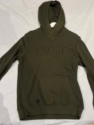 Olivgrön hoodie från Morris - Snygg olivgrön hoodie från Morris med stor magficka och broderad logga på bröstet. Klassisk huva och ribbade muddar vid ärmslut och nederkant. Perfekt för en avslappnad och trendig stil.
