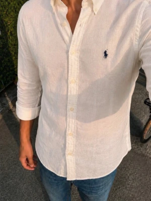 Vit linneskjorta från Ralph Lauren - Klassisk vit skjorta i linne från Ralph Lauren med den ikoniska blå broderade loggan på bröstet. Skjortan har lång ärm, knappar framtill och en avslappnad passform. Perfekt för en stilren och fräsch look.