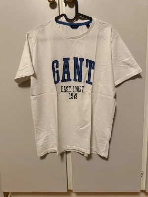 GANT t-shirt - Snygg vit t-shirt från GANT med stor blå text 'GANT' och svart text 'EAST COAST 1949' på bröstet. Klassisk rund halsringning och korta ärmar. Tillverkad i mjuk bomull som känns skön mot huden. Perfekt för en avslappnad och stilren look. Storlek 176 cm