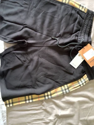 Svarta Burberry shorts - Snygga svarta shorts från Burberry med klassiska beige-rutiga detaljer längs sidorna. Shortsen har snörning i midjan och är tillverkade i mjukt bomullsmaterial. Perfekta för en avslappnad och trendig look.