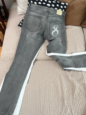 Grå jeans med vita sidor från Sterlingz Departement  - Säljer ett par grå jeans från Sterlingz Departement med raka ben och vita sidoremsor längs hela byxbenen. Jeansen har fem fickor, bälteshällor och en stor vit logga på ena bakfickan. Materialet är klassisk denim med en cool tvättad look.