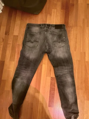 Slim fit slitna jeans från replay - Säljer ett par mörkgrå slim fit jeans från replay med slitningar och hål på framsidan för en riktigt cool och avslappnad look. Jeansen har klassiska fem fickor, dragkedjegylf och snygga detaljer på bakfickorna. Perfekta för dig som gillar streetstyle.