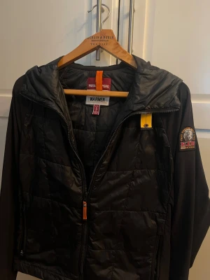 Svart cardigan från Parajumpers - Svart cardigan från Parajumpers med huva och dragkedja framtill. Jackan har en gul detalj vid halsen och ett stort patch på ärmen. Lätt vadderad och perfekt för kyligare dagar. Snygg och funktionell design.