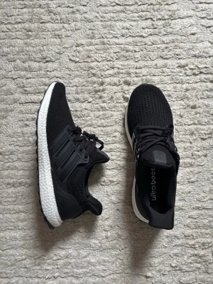 Svarta Adidas Ultraboost sneakers - Snygga svarta Adidas Ultraboost sneakers med stickad ovandel i mesh och klassiska tre ränder på sidan. Skorna har en vit, responsiv Boost-sula och svart yttersula med greppvänligt mönster. Perfekta för dig som gillar sportig och modern stil.