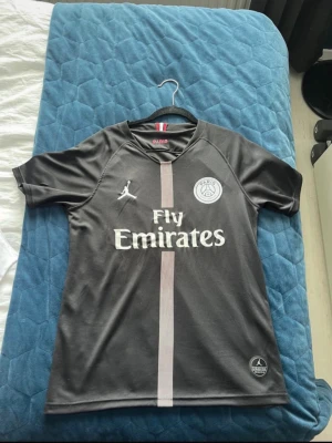 Svart PSG matchtröja Jordan x Fly Emirates - Säljer en svart PSG matchtröja i samarbete med Jordan och Fly Emirates. Tröjan har korta ärmar, rund hals och en vertikal rand i ljusgrått framtill. PSG-logga och Jordan-logga på bröstet, samt Fly Emirates-tryck. Tillverkad i lätt och snabbtorkande material.