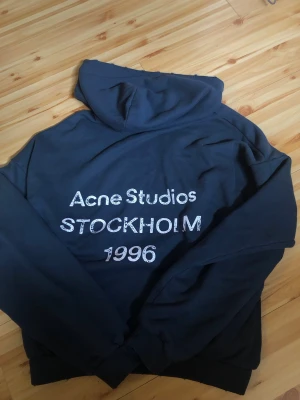 Mörkblå hoodie Acne Studios - Snygg mörkblå hoodie från Acne Studios med vit text 'Acne Studios STOCKHOLM 1996' på framsidan. Klassisk passform med huva och ribbade muddar. Perfekt för dig som gillar streetwear och vill ha en stilren look.