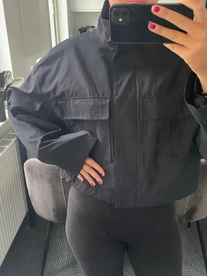 Svart kort bomberjacka med fickor - Säljer en svart bomberjacka med hög krage och stora framfickor. Jackan har dragkedja framtill och en croppad, loose passform. Perfekt för dig som gillar minimalistisk stil och vill ha en enkel men snygg jacka till vardags.   Mörk mörk blå är dne nästan svart 