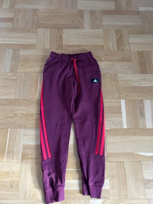 Vinröda Adidas mjukisbyxor med röda ränder - Säljer ett par vinröda mjukisbyxor från Adidas med klassiska röda ränder längs benen och en liten Adidas-logga på vänster sida. Byxorna har resår i midjan med röd dragsko och muddar vid bensluten. Perfekta för chill eller träning. Har matchande hoodie på min profil:)
