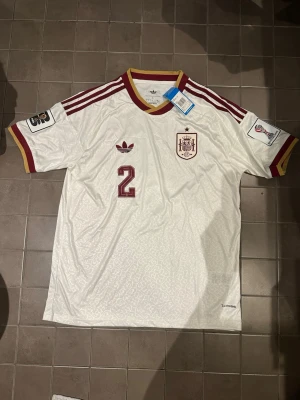 Vit Spanien Adidas fotbollströja - Säljer en vit Spanien fotbollströja från Adidas med röda detaljer och nummer 2 på bröstet. Tröjan har gul och röd krage, röda ränder på axlarna och officiella märken på ärmarna. Perfekt för dig som älskar fotboll och vill stötta Spanien.