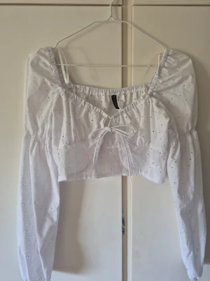 Vit croppad blus med knyt - Söt vit croppad blus från H&M med broderade detaljer och knyt framtill. Blusen har lång ärm och elastisk kant vid axlarna för en offshoulder-look. Perfekt till sommaren och ger en romantisk vibe. Går att ha över ett linne, bli super fint. Säljer då den inte används, har bara använt den 1 gång!. Köpt för 150 kr