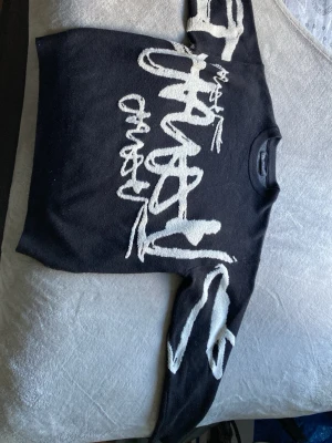 Svart stickad tröja med vit text - Cool svart stickad tröja med stor vit grafisk text över hela framsidan och ärmarna. Tröjan har rund halsringning och avslappnad passform. Perfekt för dig som gillar streetwear och vill sticka ut lite extra.