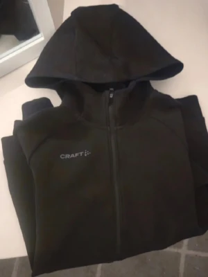 Svart huvtröja från craft  - Svart hoodie från Craft med hel dragkedja framtill och två fickor med dragkedja. Hoodien har en stilren design med logga på bröstet och ribbade muddar vid ärmslut och nederkant. Perfekt för en sportig och avslappnad look.