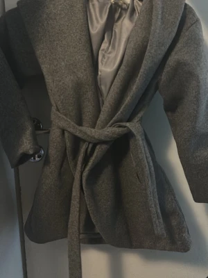 Grå kappa med knytskärp - Stilren grå kappa med bred krage och knytskärp i midjan. Kappan har slät yta, är helfodrad och har långa ärmar. Perfekt för kyligare dagar och ger en klassisk look till din outfit.