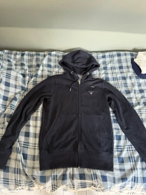 Mörkblå hoodie från Gant - Säljer en mörkblå hoodie från Gant med dragkedja framtill och två stora fickor. Klassisk huva med dragsko och Gant-logga broderad på bröstet. Tillverkad i mjukt bomullsmaterial som är skönt att ha på sig. Perfekt för en avslappnad och stilren look.