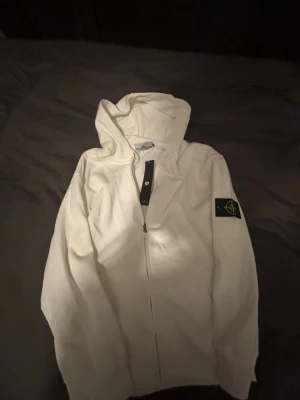 Vit hoodie från Stone Island - Snygg vit hoodie från Stone Island med dragkedja framtill och klassisk svart patch med gult kompassmärke på vänster ärm. Hoodien har huva och en clean look som passar perfekt till streetwear-stilen. Materialet känns mjukt och skönt.