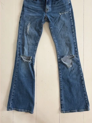 Blå slitna bootcut jeans - Säljer ett par blå bootcut jeans med slitningar och trasiga detaljer på benen. Jeansen har klassisk femficksdesign och är tillverkade i denim. Perfekt för dig som gillar en avslappnad och trendig look.
