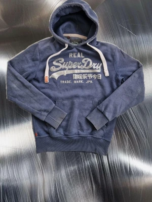 Blå Superdry hoodie - Snygg blå hoodie från Superdry med stort silvrigt tryck på bröstet och japanska tecken. Liten fläck som knappt syns finns på ena armen (se bild 6), går mest troligt bort efter en tvätt. Mått: Armlängd: 65cm armhåla-armhåla (bredd) : 52cm Armbredd: 15 cm axel-midejslut (längd) : 65cm