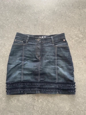 Mörk jeanskjol med volang från Cotonel - Snygg mörkblå jeanskjol från Cotonel med volangdetaljer längst ner och små rosa hjärtan broderade på insidan av linningen. Kjolen har klassisk femficksdesign och knappstängning framtill. Perfekt för dig som gillar en söt men cool stil.