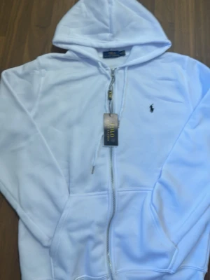 Polo ralph lauren - Zip up hoodie - Snygg vit hoodie från Polo Ralph Lauren med dragkedja framtill och klassisk huva. Märkets ikoniska logga är broderad på bröstet. Hoodien har dragsko i huvan och är tillverkad i mjukt material som känns skönt mot huden. Perfekt för en clean och stilren look. Den enda defekten är tag där bak på "P" av polo ralph lauren