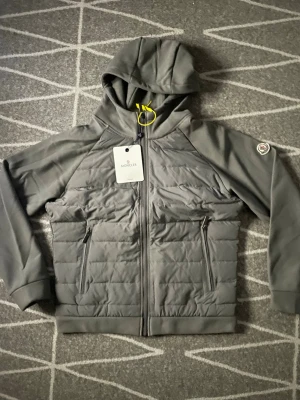 Grå pufferjacka från Moncler - Snygg grå pufferjacka från Moncler med huva och dragkedja framtill. Jackan har två praktiska fickor med dragkedja och Moncler-logga på ärmen. Tillverkad i ett quiltat material som håller värmen under kalla dagar.