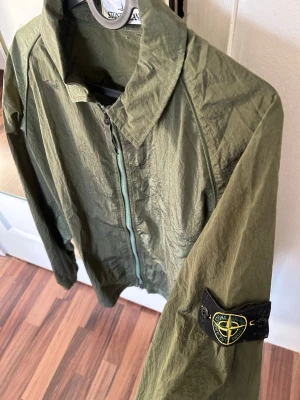 Olivgrön vindjacka från Stone Island - Snygg olivgrön vindjacka från Stone Island med klassisk patch på ärmen. Jackan har dragkedja framtill, ståkrage och är tillverkad i ett lätt, glansigt material som ger en sportig look. Perfekt för dig som gillar streetwear och vill ha något stilrent. Köpt här på Plick