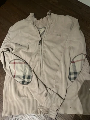 Beige ziptröja från Burberry Brit - Säljer en beige ziptröja från Burberry Brit med ribbade muddar och klassiska rutiga armbågslappar. Tröjan har hög krage och dragkedja framtill. Perfekt för dig som gillar stilrena detaljer och exklusiva märken.