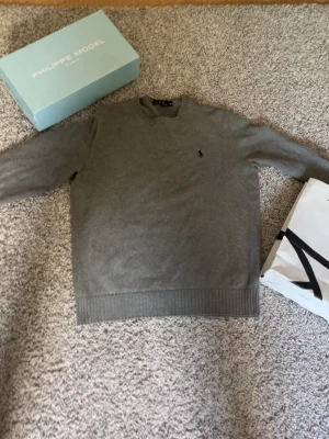 Grå stickad tröja Polo Ralph Lauren - Snygg grå stickad tröja från Polo Ralph Lauren med rund halsringning och diskret broderad logga på bröstet. Tröjan har ribbade muddar vid ärmslut och nederkant. Perfekt till lager på lager och enkel att matcha med jeans eller chinos.