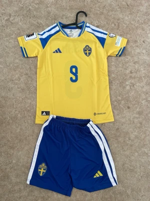 Sverige fotbollströja och shorts Adidas - Säljer ett matchställ från svenska landslaget med gul tröja och blå shorts. Tröjan har nummer 9 och namnet ISAK på ryggen, samt blå detaljer och landslagets emblem. Shortsen är blå med vita ränder och har också emblemet. Perfekt för fotbollsträning eller match.