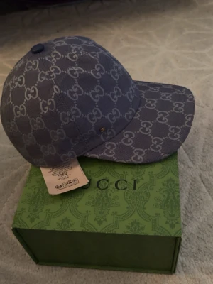 Blå GG-monogram keps från Gucci - Säljer en blå keps från Gucci med det klassiska GG-monogrammet över hela kepsen. Kepsen har böjd skärm, justerbart band bak i blått läder och är tillverkad i canvasmaterial. Kommer med original grön Gucci-box. Perfekt för dig som gillar exklusiva accessoarer.