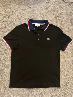 Svart pikétröja från Lacoste - Klassisk svart pikétröja från Lacoste med blå krage och vita samt röda detaljer. Tröjan har korta ärmar med matchande kantband och den ikoniska gröna krokodilloggan på bröstet. Perfekt till jeans eller shorts för en clean look.