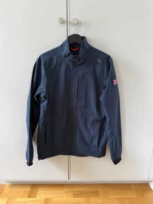 Mörkblå vindjacka från Sail Racing - Snygg mörkblå vindjacka från Sail Racing med hög krage och hel dragkedja framtill. Jackan har en bröstficka med dragkedja, två sidofickor och en Union Jack-patch på ena ärmen. Tillverkad i ett lätt och vindtätt material.