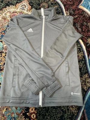 Grå träningsjacka från Adidas - Snygg grå träningsjacka från Adidas med vit dragkedja och hög krage. Jackan har två sidofickor, elastiska muddar vid ärmsluten och AEROREADY-tryck på framsidan. Perfekt för träning eller chill. Tillverkad i ett mjukt funktionsmaterial.