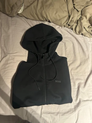 Svart zip hoodie från BOSS - Snygg svart hoodie från BOSS med dragkedja framtill och justerbar huva med snören. Diskret BOSS-logga broderad på bröstet. Hoodien har två fickor på sidorna och är tillverkad i ett mjukt material som känns skönt mot huden.
