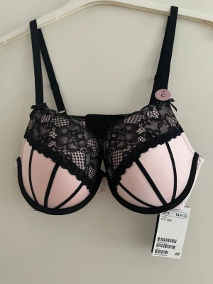 Rosa och svart spets-bh från H&M - Snygg bh i ljusrosa med svarta detaljer och spets från H&M. Kuporna har dekorativa svarta band och överdelen är täckt av blommig spets. Justerbara axelband och hakknäppning bak. Perfekt för dig som gillar en feminin och trendig look.