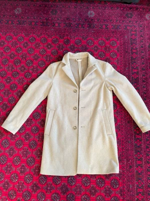 Beige ullkappa från H&M - Stilren beige kappa från H&M med klassisk krage och tre knappar framtill. Kappan har raka fickor på sidorna och är tillverkad i en ullblandning som ger en mjuk och varm känsla. Perfekt för kyliga dagar och enkel att matcha med olika outfits.