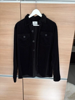Svart manchester overshirt från Ciszere - Snygg svart overshirt i manchestertyg från Ciszere. Skjortan har klassisk krage, knäppning framtill och två bröstfickor med lock och knapp. Perfekt lager på lager-plagg med avslappnad passform och långa ärmar.