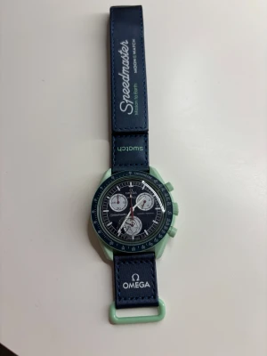Omega x Swatch Speedmaster klocka - Säljer en Omega x Swatch Speedmaster MoonSwatch Mission on Earth. Klockan har blått band med vita loggor och mintgrönt boett. Snygg sportig look med flera urtavlor och tydliga detaljer. Perfekt för dig som gillar coola accessoarer och ikonisk design.