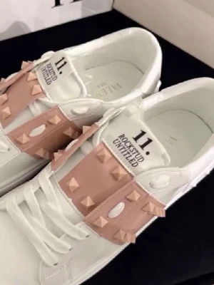 Valentino Rockstud Untitled sneakers - Snygga vita sneakers från Valentino med ljusrosa band och ikoniska pyramidnitar. Skorna har en låg siluett, rund tå och är tillverkade i läder. Perfekta för dig som vill ha en unik och trendig look med exklusiva detaljer.